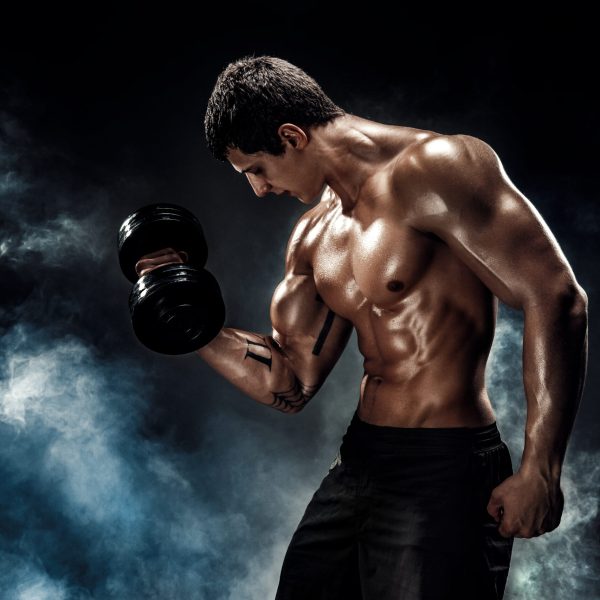 Handsome bodybuilder doing exercise on biceps with dumbbell. Studio shot. Smoke.